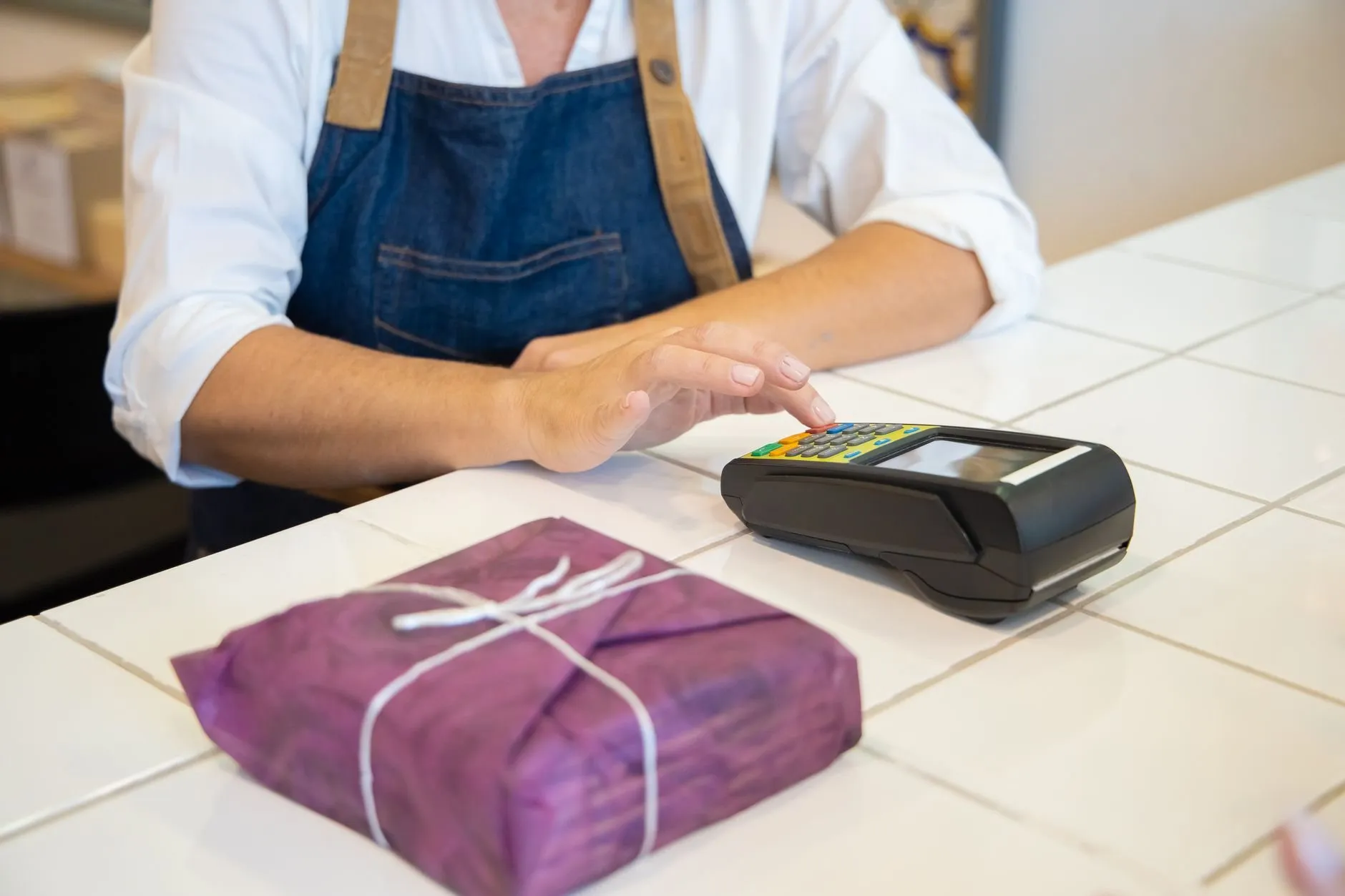 A person in a denim apron at a tiled counter with a card terminal and a wrapped parcel