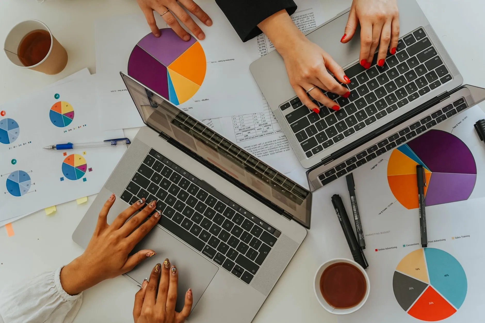 Four hands at two laptops on a white desk covered in printed pie charts
