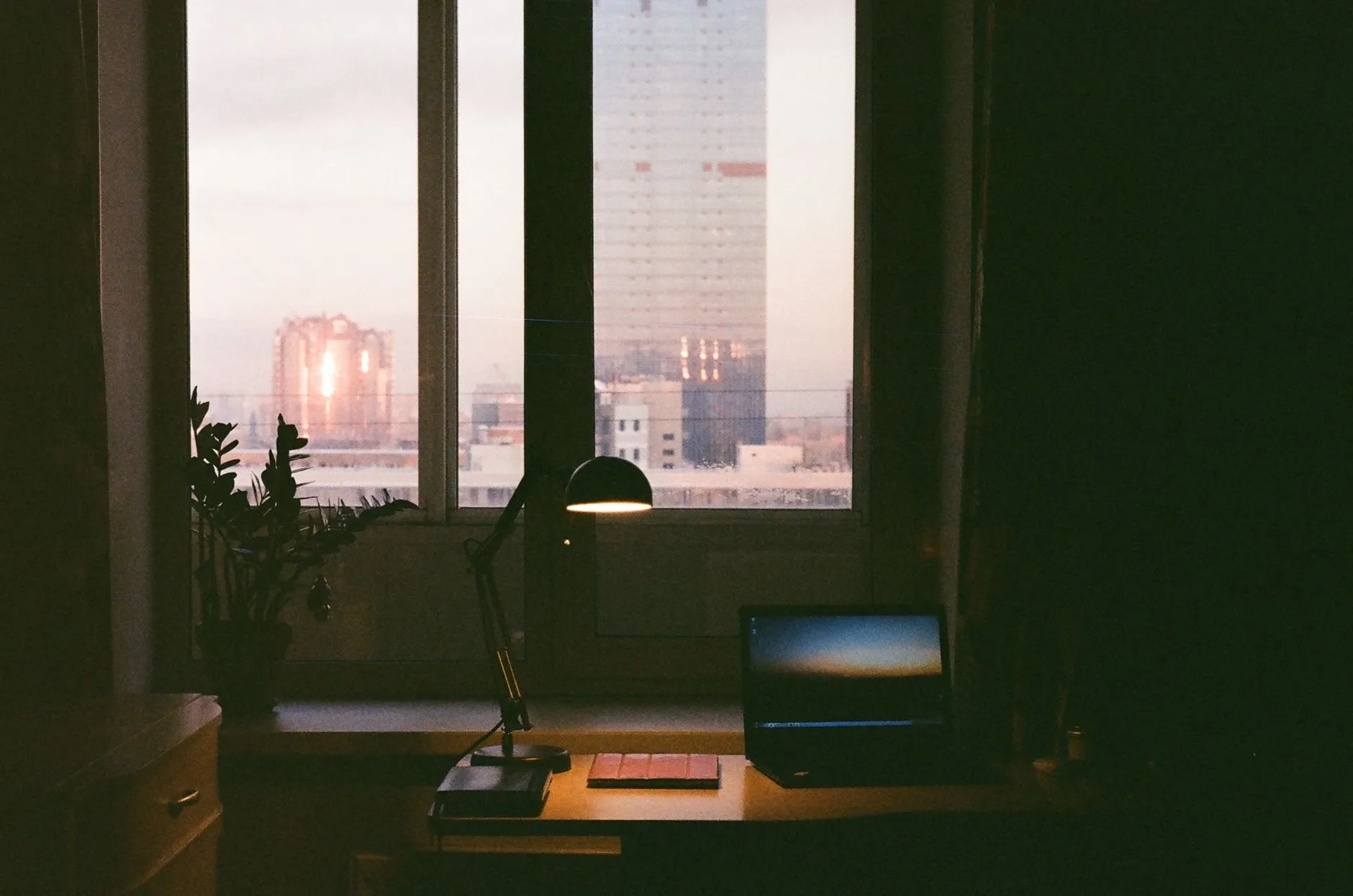 A desk lamp lit over a laptop by a window at dusk, city towers outside