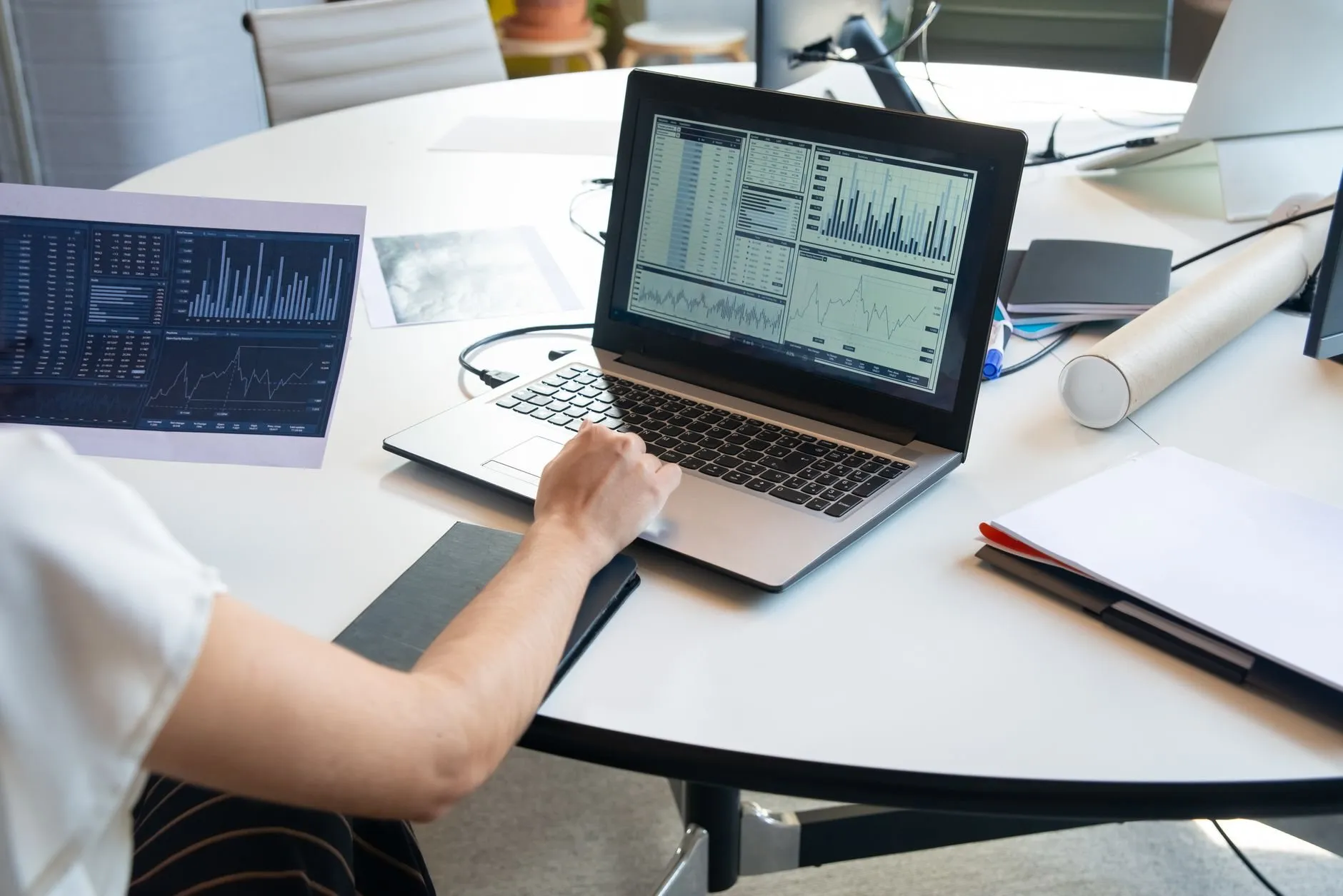 A person's arm at a laptop showing bar and line charts on a round white meeting table