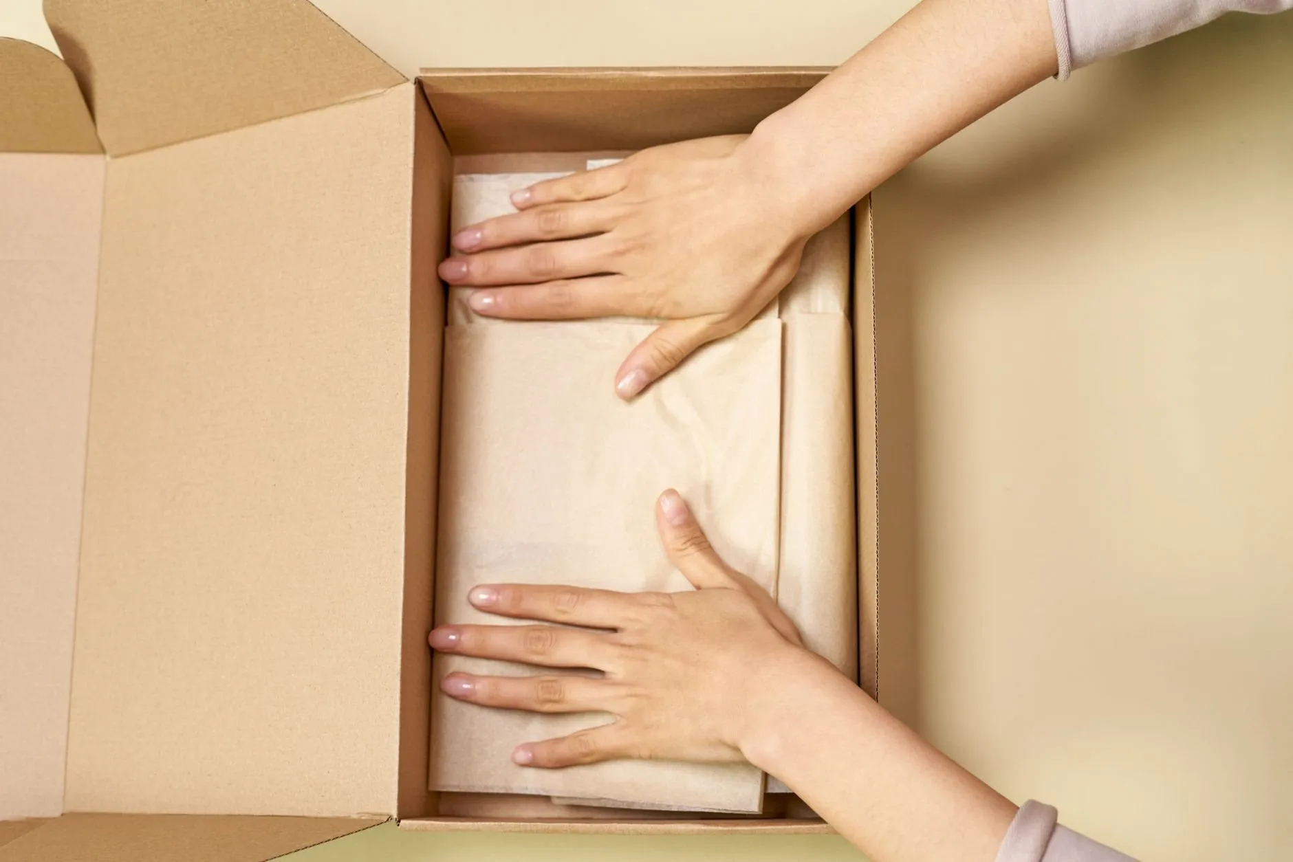 Two hands smoothing tissue paper inside an open cardboard box on a pale table